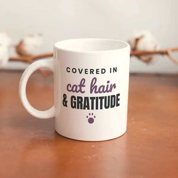 White 11 oz ceramic mug printed with "Covered in Cat Hair and Gratitude" on a wooden table