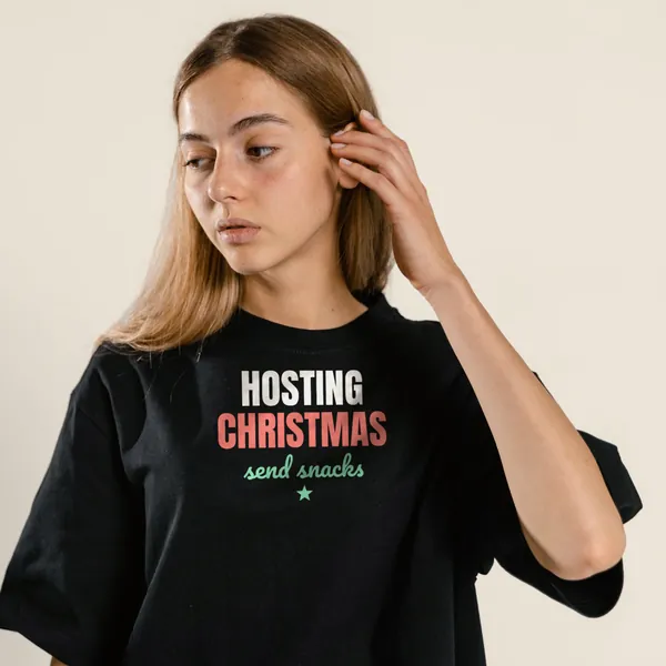 Model wearing a black "Hosting Christmas, Send Snacks" tee with the light-ink version of the design